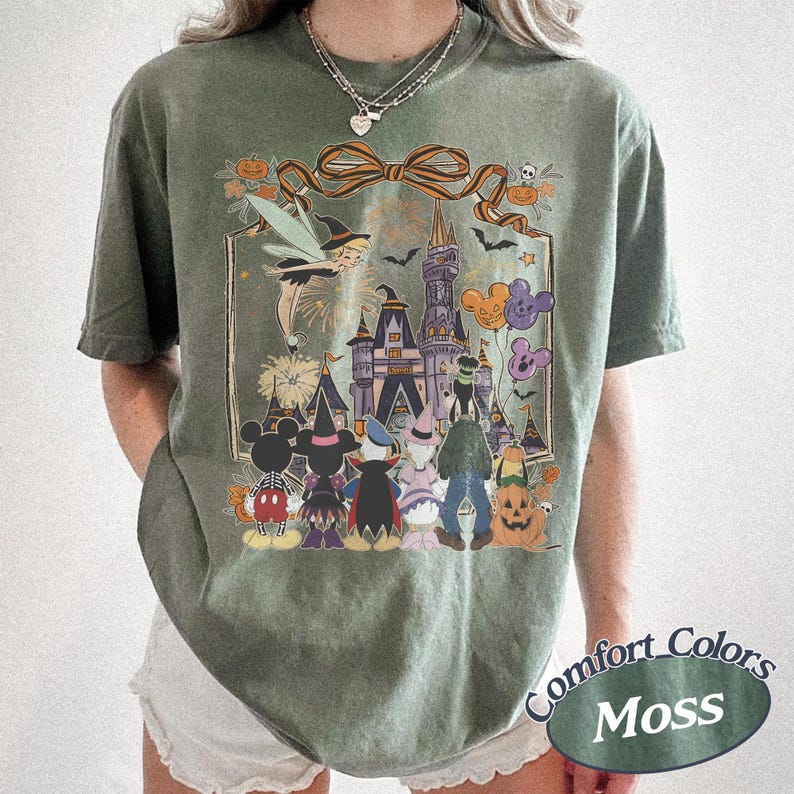 Pode incluir: T-shirt verde musgo com um gr&aacute;fico de Halloween com um castelo, personagens e ab&oacute;boras. O design inclui uma bruxa, fogos de artif&iacute;cio e bal&otilde;es em forma de Mickey Mouse. A camisa tem uma etiqueta Comfort Colors.