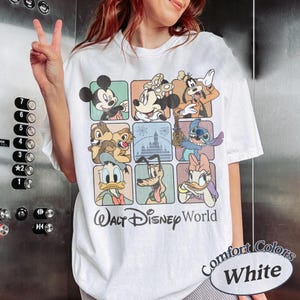 May include: White t-shirt featuring a grid of Disney characters including Mickey Mouse, Minnie Mouse, and Goofy. The shirt has the text "Walt Disney World" and "Comfort Colors White" printed on it. The shirt is a short-sleeved, casual style.
