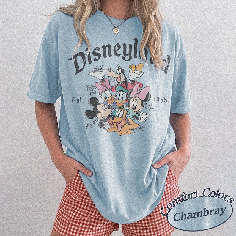 May include: Light blue t-shirt with a graphic of Disney characters, including Mickey Mouse, Minnie Mouse, Donald Duck, and Goofy. The shirt has the text "Disneyland" and "Est. 1955." The shirt is made of Comfort Colors Chambray material.