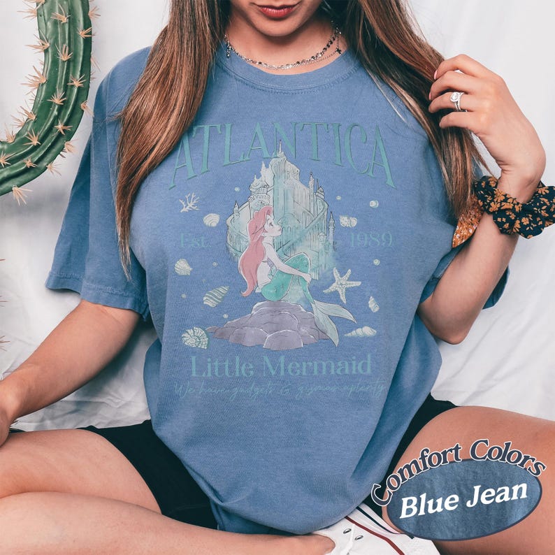 Może przedstawiać: Niebieska koszulka w kolorze dżinsu z grafiką syreny i zamku z napisami "Atlantica" i "Little Mermaid". Koszulka ma wygląd vintage i postarzaną fakturę, z napisem "Comfort Colors" wydrukowanym w prawym dolnym rogu.
