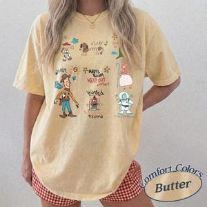 Puede incluir: Camiseta de color mantequilla con personajes de dibujos animados y texto. El dise&ntilde;o incluye a Woody, Slinky y Buzz Lightyear, con las palabras "ANDYS ROOM KEEP OUT" y "WANTED REWARD". La camiseta es de la marca Comfort Colors.