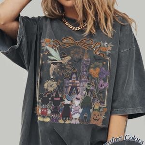 Pode incluir: Camiseta cinza escuro Comfort Colors com um gr&aacute;fico tem&aacute;tico de Halloween. O design inclui um castelo, fogos de artif&iacute;cio e personagens de desenhos animados fantasiados. O texto "Comfort Colors Pepper" &eacute; impresso no canto inferior direito.