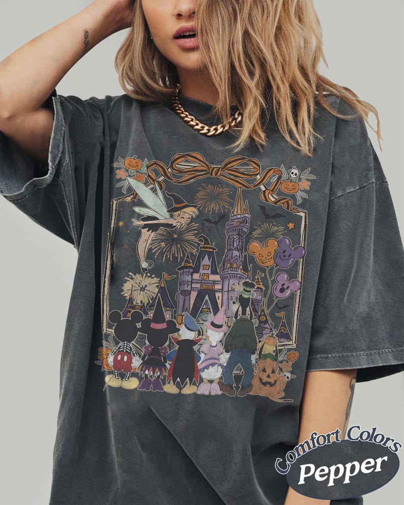 Pode incluir: Camiseta cinza escuro Comfort Colors com um gr&aacute;fico tem&aacute;tico de Halloween. O design inclui um castelo, fogos de artif&iacute;cio e personagens de desenhos animados fantasiados. O texto "Comfort Colors Pepper" &eacute; impresso no canto inferior direito.