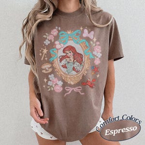 Puede incluir: Camiseta marr&oacute;n con una ilustraci&oacute;n enmarcada de una princesa pelirroja, lazos turquesa y motivos florales y n&aacute;uticos. La camiseta tiene las palabras "Comfort Colors Espresso" impresas en la parte inferior derecha.