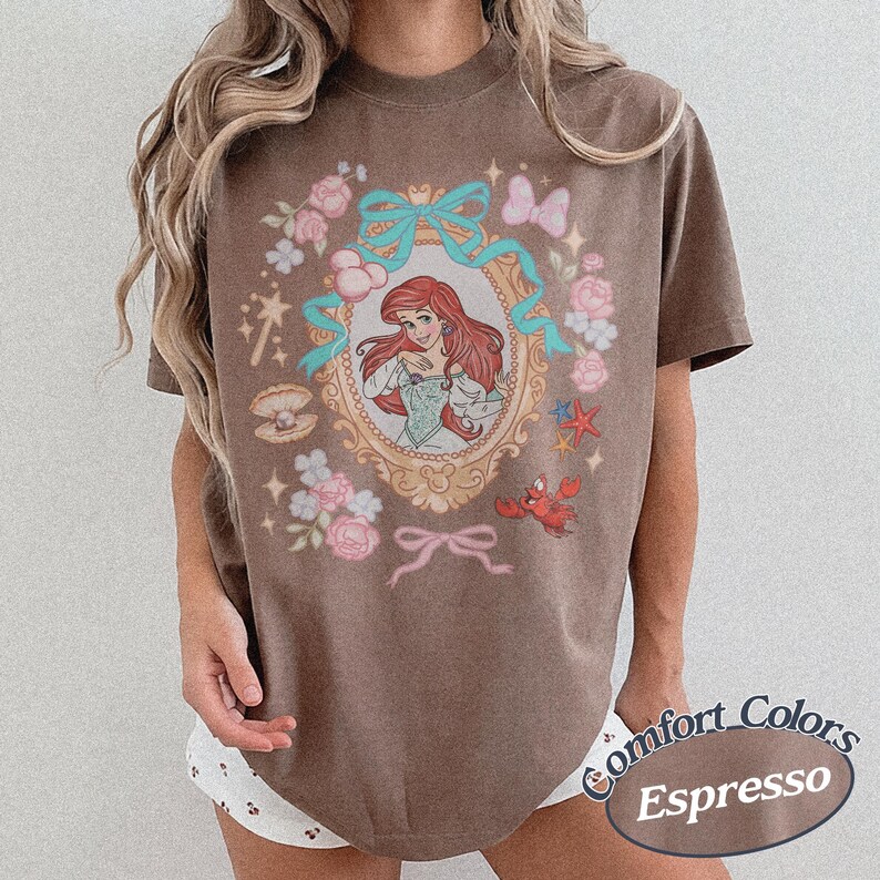 Puede incluir: Camiseta marr&oacute;n con una ilustraci&oacute;n enmarcada de una princesa pelirroja, lazos turquesa y motivos florales y n&aacute;uticos. La camiseta tiene las palabras "Comfort Colors Espresso" impresas en la parte inferior derecha.