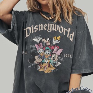 Może przedstawiać: Ciemnoszara koszulka z nadrukiem "Disneyworld" z Myszką Miki, Myszka Minnie, Kaczorem Donaldem, Daisy, Goofym i Pluto. Koszulka zawiera tekst "Est. 1971" i "Comfort Colors Pepper".