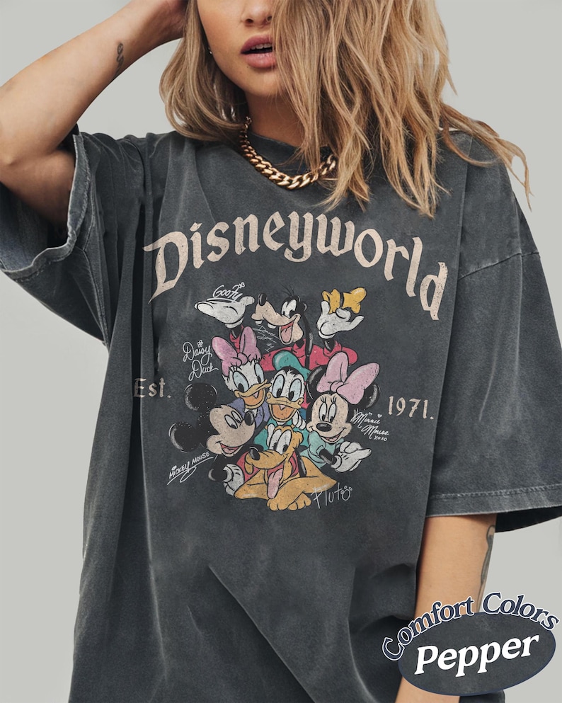 Może przedstawiać: Ciemnoszara koszulka z nadrukiem "Disneyworld" z Myszką Miki, Myszka Minnie, Kaczorem Donaldem, Daisy, Goofym i Pluto. Koszulka zawiera tekst "Est. 1971" i "Comfort Colors Pepper".