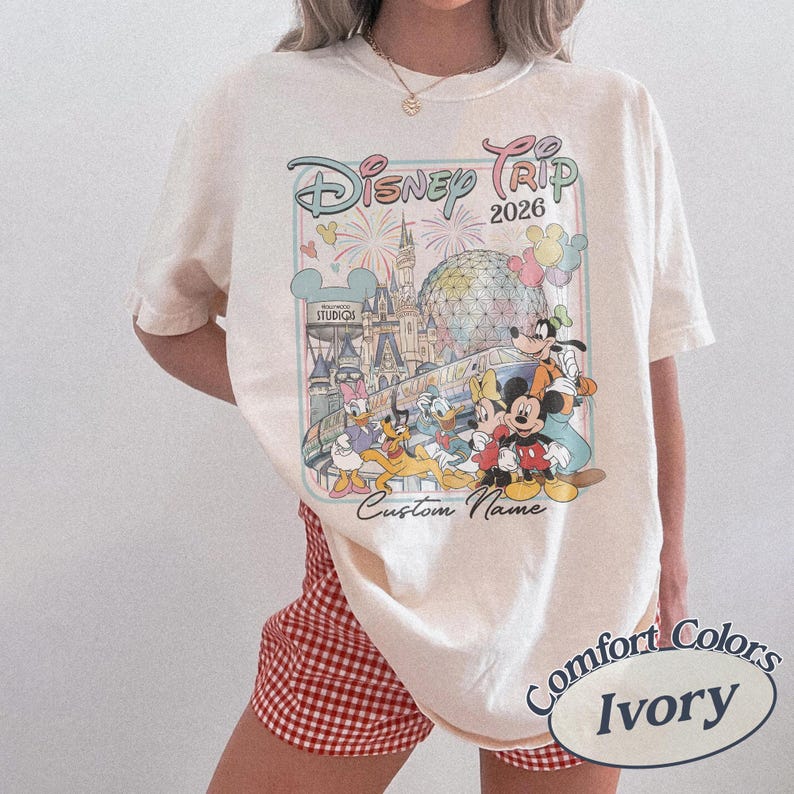 May include: Cream-coloured t-shirt with a Disney Trip 2026 graphic featuring Mickey Mouse and friends. The design includes a castle and fireworks. The shirt has a crew neck and short sleeves. The words "Custom Name" are printed below the graphic.