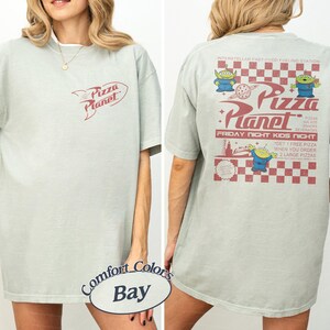May include: A light green t-shirt with the Pizza Planet logo on the front and back. The back of the shirt features the Pizza Planet logo, alien characters, and text that reads "Friday Night Kids Night."
