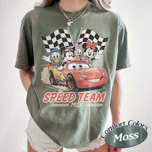 K&ouml;nnte beinhalten: Moosgr&uuml;nes T-Shirt mit einer Grafik von Disney-Figuren in einem roten Rennwagen und Schachflaggen. Der Text "SPEED TEAM 1928" ist unter dem Auto aufgedruckt. Das T-Shirt ist von der Marke Comfort Colors.