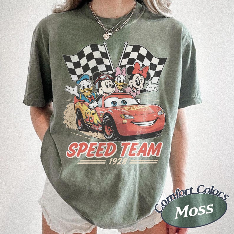 K&ouml;nnte beinhalten: Moosgr&uuml;nes T-Shirt mit einer Grafik von Disney-Figuren in einem roten Rennwagen und Schachflaggen. Der Text "SPEED TEAM 1928" ist unter dem Auto aufgedruckt. Das T-Shirt ist von der Marke Comfort Colors.