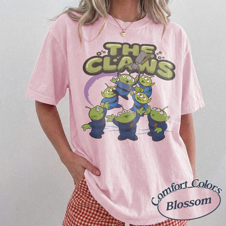 May include: Pink t-shirt featuring a graphic of green aliens in blue uniforms with the text "THE CLAWS" above them. The shirt has a Comfort Colors Blossom label.