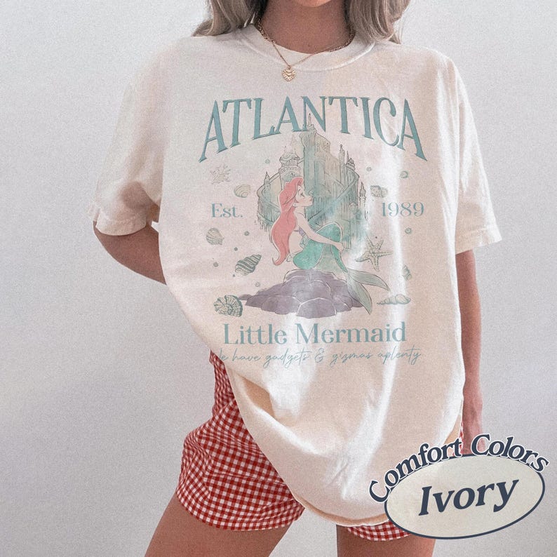 Może przedstawiać: Kremowa koszulka z nadrukiem z napisem "ATLANTICA" i "Little Mermaid". Projekt zawiera ilustrację syreny, zamek i tekst "Est. 1989". Koszulka jest częścią kolekcji Comfort Colors.