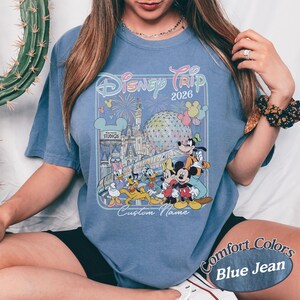 May include: Blue jean coloured t-shirt with a colourful graphic of Disney characters, fireworks, and the text "Disney Trip 2026". The shirt also has the text "Custom Name" and "Comfort Colors".