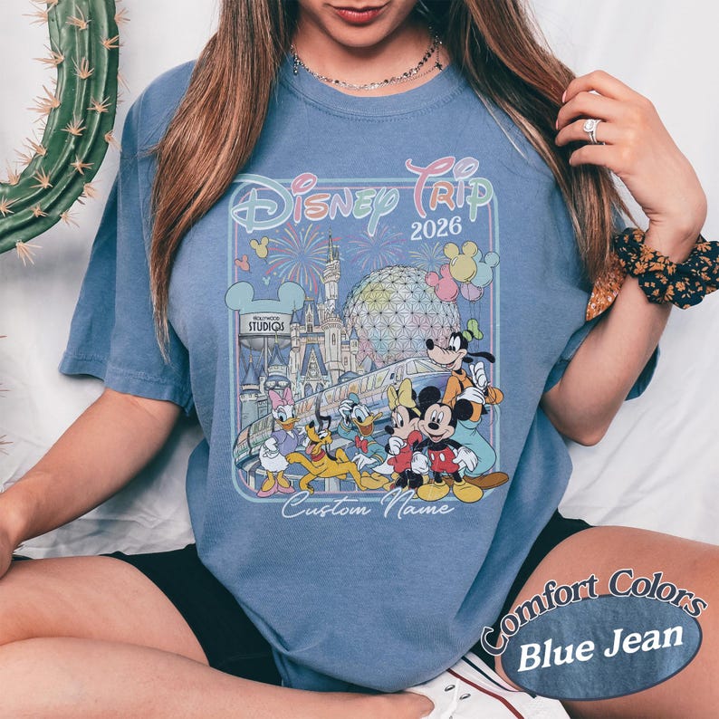 May include: Blue jean coloured t-shirt with a colourful graphic of Disney characters, fireworks, and the text "Disney Trip 2026". The shirt also has the text "Custom Name" and "Comfort Colors".