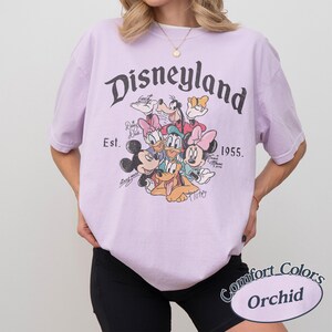 May include: Orchid-colored t-shirt featuring a Disneyland graphic with Mickey Mouse, Minnie Mouse, Donald Duck, Daisy Duck, Goofy, and Pluto. The shirt includes the text "Disneyland" and "Est. 1955."
