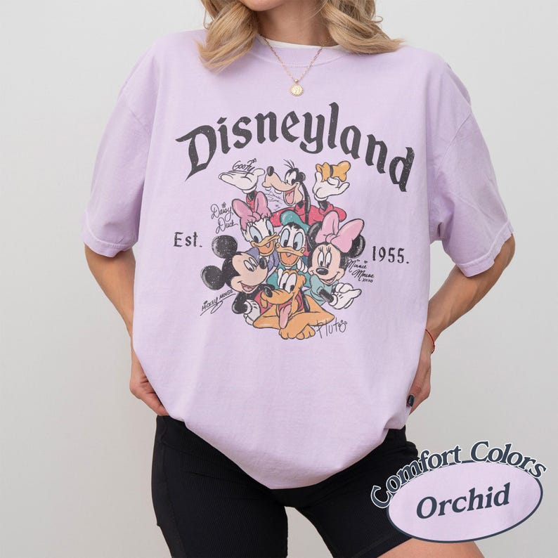 May include: Orchid-colored t-shirt featuring a Disneyland graphic with Mickey Mouse, Minnie Mouse, Donald Duck, Daisy Duck, Goofy, and Pluto. The shirt includes the text "Disneyland" and "Est. 1955."