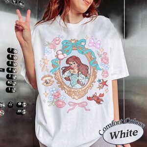 Puede incluir: Camiseta blanca con una ilustraci&oacute;n enmarcada de una sirena con pelo rojo, rodeada de rosas rosas, lazos azules y elementos marinos. La camiseta tiene la inscripci&oacute;n "Comfort Colors White".