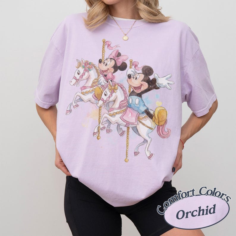 Puede incluir: Camiseta lila claro con un dise&ntilde;o de carrusel con Mickey y Minnie Mouse montados en caballos blancos. El gr&aacute;fico tiene un efecto de acuarela. La camiseta est&aacute; etiquetada como "Comfort Colors" y "Orchid" en un &oacute;valo blanco.