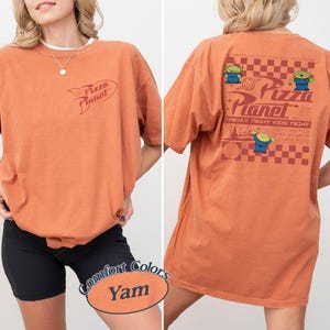 May include: Orange-colored t-shirt with the Pizza Planet logo on the front and back. The back of the shirt features the Pizza Planet logo, three green alien characters, and text that reads "Friday Night Kids Night".