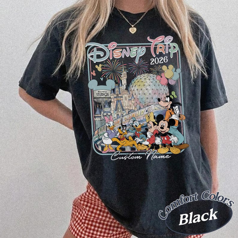 May include: Black t-shirt with a Disney Trip 2026 design, showcasing Mickey Mouse, Donald Duck, and friends. The graphic includes fireworks, a castle, and "Custom Name". The shirt is labelled "Comfort Colors Black".
