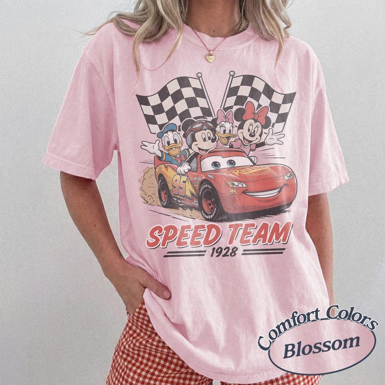 K&ouml;nnte beinhalten: Hellrosa T-Shirt mit einem Aufdruck von Disney-Figuren in einem Rennwagen mit dem Text "SPEED TEAM 1928". Das Design umfasst Micky Maus, Donald Duck, Daisy Duck und Minnie Maus, mit karierten Flaggen. Das Shirt ist ein Comfort Colors Blossom.