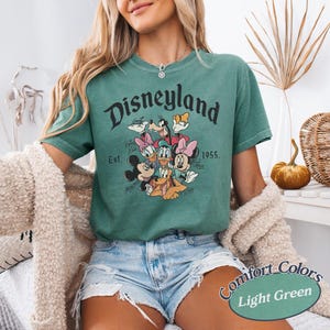 May include: Light green t-shirt featuring the word "Disneyland" above a graphic of Disney characters. The shirt includes the text "Est. 1955." and is labeled "Comfort Colors Light Green".