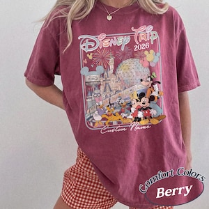 May include: A berry-coloured t-shirt with a Disney Trip 2026 graphic, showcasing Disney characters and park scenes. The design includes fireworks, a castle, and "Custom Name". The shirt is labelled "Comfort Colors".