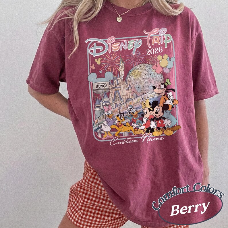 May include: A berry-coloured t-shirt with a Disney Trip 2026 graphic, showcasing Disney characters and park scenes. The design includes fireworks, a castle, and "Custom Name". The shirt is labelled "Comfort Colors".