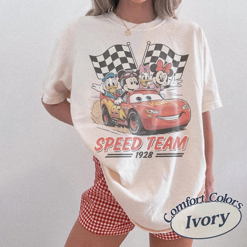 K&ouml;nnte beinhalten: Elfenbeinfarbenes T-Shirt mit einem Aufdruck von Mickey Mouse, Donald Duck, Daisy Duck und Minnie Mouse in einem roten Rennwagen mit dem Text "Speed Team 1928". Zwei karierte Flaggen im Hintergrund.