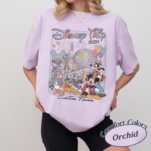 May include: A lilac t-shirt with a Disney-themed graphic, showcasing Mickey Mouse, Donald Duck, and iconic landmarks. The design includes the text "Disney Trip 2026" and "Custom Name". The shirt is a Comfort Colors Orchid.