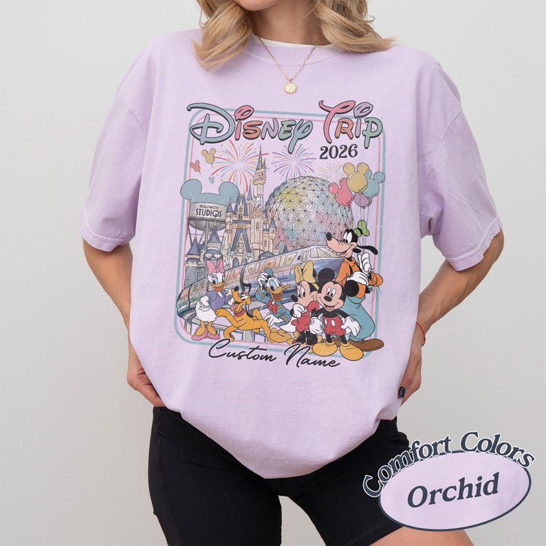 May include: A lilac t-shirt with a Disney-themed graphic, showcasing Mickey Mouse, Donald Duck, and iconic landmarks. The design includes the text "Disney Trip 2026" and "Custom Name". The shirt is a Comfort Colors Orchid.
