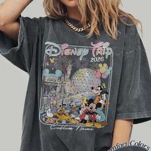 May include: Dark grey t-shirt with a Disney Trip 2026 graphic, showcasing Disney characters, fireworks, and a castle. The shirt includes the text "Custom Name" and "Comfort Colors Pepper".