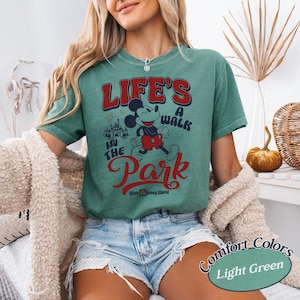 May include: Light green t-shirt with a graphic of Mickey Mouse and the text "LIFE'S A WALK IN THE PARK" in red and white. The shirt also features the words "Walt Disney World" in white. The shirt is a Comfort Colors brand.