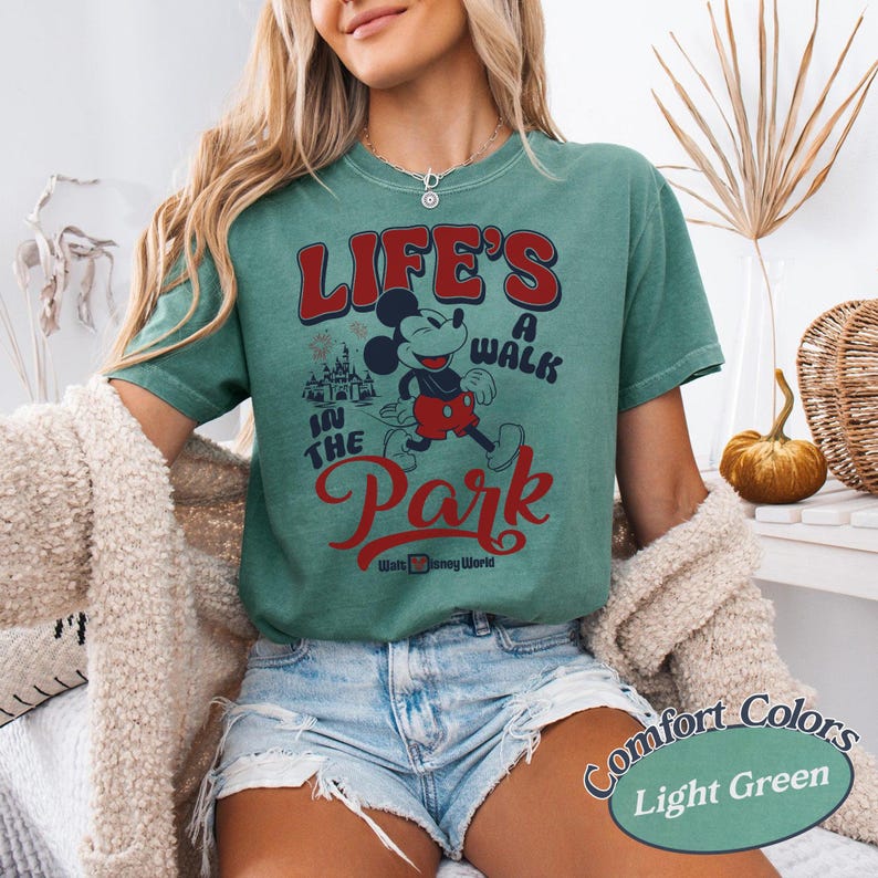 May include: Light green t-shirt with a graphic of Mickey Mouse and the text "LIFE'S A WALK IN THE PARK" in red and white. The shirt also features the words "Walt Disney World" in white. The shirt is a Comfort Colors brand.