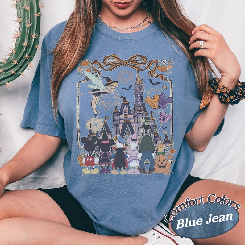 Pode incluir: Camiseta azul jeans Comfort Colors com um gr&aacute;fico de Halloween. O gr&aacute;fico inclui um castelo, personagens e o texto "Comfort Colors Blue Jean". O design incorpora uma fada, ab&oacute;boras e morcegos.