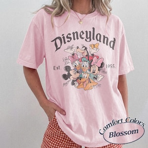 May include: A light pink t-shirt featuring the word "Disneyland" in a vintage font, along with a graphic of Disney characters. The shirt includes the text "Est. 1955" and "Comfort Colors Blossom".