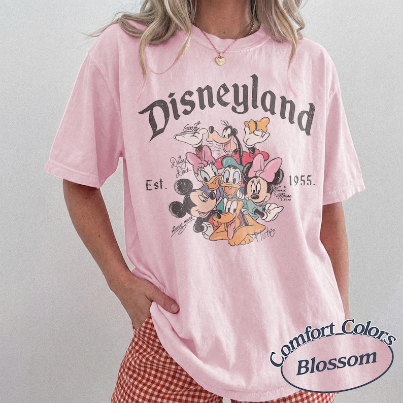 May include: A light pink t-shirt featuring the word "Disneyland" in a vintage font, along with a graphic of Disney characters. The shirt includes the text "Est. 1955" and "Comfort Colors Blossom".
