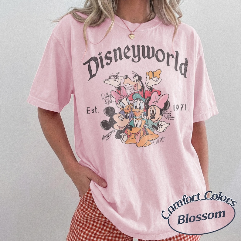 Może przedstawiać: Jasnor&oacute;żowa koszulka z napisem "Disneyworld" i grafiką postaci Disneya, w tym Myszki Miki i Kaczora Donalda. Koszulka zawiera r&oacute;wnież tekst "Est. 1971" i "Comfort Colors Blossom".