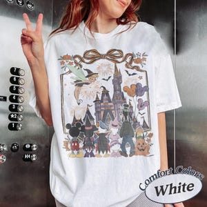 Pode incluir: Camiseta branca Comfort Colors com um gr&aacute;fico tem&aacute;tico de Halloween. O design inclui um castelo, personagens, fogos de artif&iacute;cio e bal&otilde;es do Mickey Mouse. A camiseta tem gola redonda e mangas curtas.