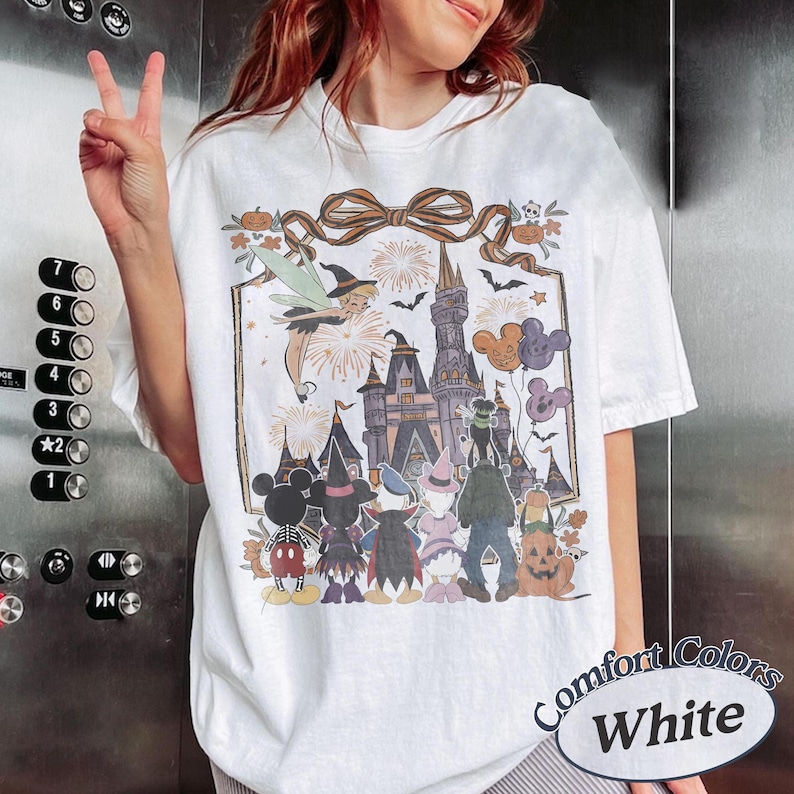 Pode incluir: Camiseta branca Comfort Colors com um gr&aacute;fico tem&aacute;tico de Halloween. O design inclui um castelo, personagens, fogos de artif&iacute;cio e bal&otilde;es do Mickey Mouse. A camiseta tem gola redonda e mangas curtas.