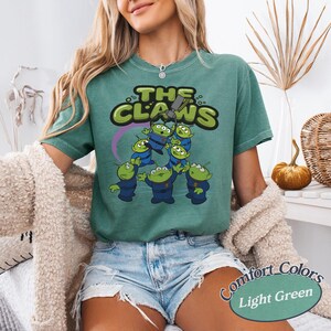 May include: Light green t-shirt featuring a graphic of the aliens from the movie Toy Story. The graphic includes the text "THE CLAWS" in green and yellow, with the aliens in blue uniforms. The shirt is labeled "Comfort Colors Light Green".
