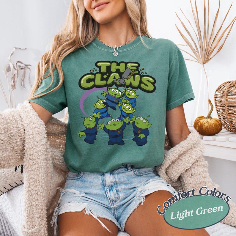 May include: Light green t-shirt featuring a graphic of the aliens from the movie Toy Story. The graphic includes the text "THE CLAWS" in green and yellow, with the aliens in blue uniforms. The shirt is labeled "Comfort Colors Light Green".