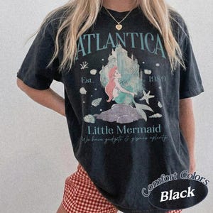 Może przedstawiać: Czarny t-shirt z grafiką z napisem "ATLANTICA" nad ilustracją syreny i zamku. Koszulka zawiera r&oacute;wnież napis "Little Mermaid" i "1989".