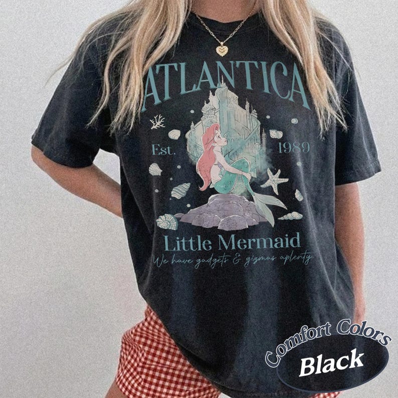 Może przedstawiać: Czarny t-shirt z grafiką z napisem "ATLANTICA" nad ilustracją syreny i zamku. Koszulka zawiera r&oacute;wnież napis "Little Mermaid" i "1989".