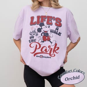 May include: Orchid-colored t-shirt featuring a graphic of Mickey Mouse walking with the text "Life's a Walk in the Park" in red and black. The shirt also includes the Walt Disney World logo.