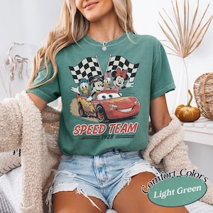 K&ouml;nnte beinhalten: Hellgr&uuml;nes T-Shirt mit einem roten Rennwagen mit Mickey, Minnie, Donald und Daisy Duck. Das Auto ist mit "95" und "Speed Team 1928" beschriftet. Zwei karierte Flaggen sind im Hintergrund. Das T-Shirt ist mit "Comfort Colors Light Green" beschriftet.