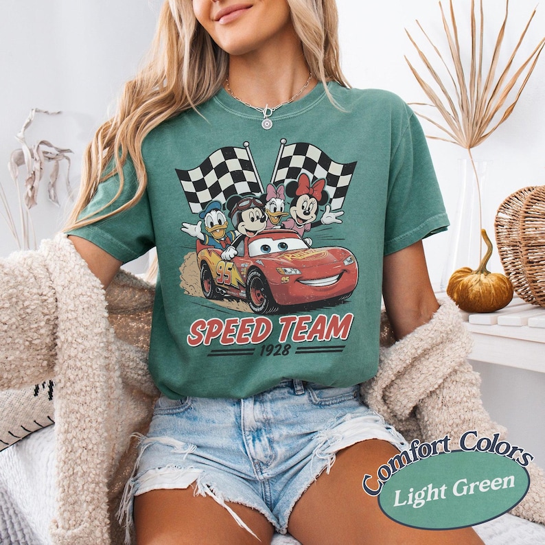 K&ouml;nnte beinhalten: Hellgr&uuml;nes T-Shirt mit einem roten Rennwagen mit Mickey, Minnie, Donald und Daisy Duck. Das Auto ist mit "95" und "Speed Team 1928" beschriftet. Zwei karierte Flaggen sind im Hintergrund. Das T-Shirt ist mit "Comfort Colors Light Green" beschriftet.