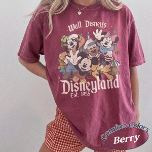 May include: Berry-colored t-shirt featuring a graphic of Disney characters, including Mickey Mouse, Donald Duck, and Minnie Mouse, with the text "Walt Disney's Disneyland Est. 1955." The shirt is a Comfort Colors brand.