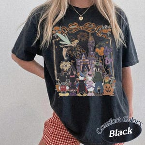 Pode incluir: Camiseta preta Comfort Colors com um gr&aacute;fico tem&aacute;tico de Halloween. O design inclui um castelo, fogos de artif&iacute;cio e personagens de desenhos animados fantasiados. A camisa tem um visual desgastado e as palavras "Comfort Colors Black" s&atilde;o impressas no canto inferior direito.
